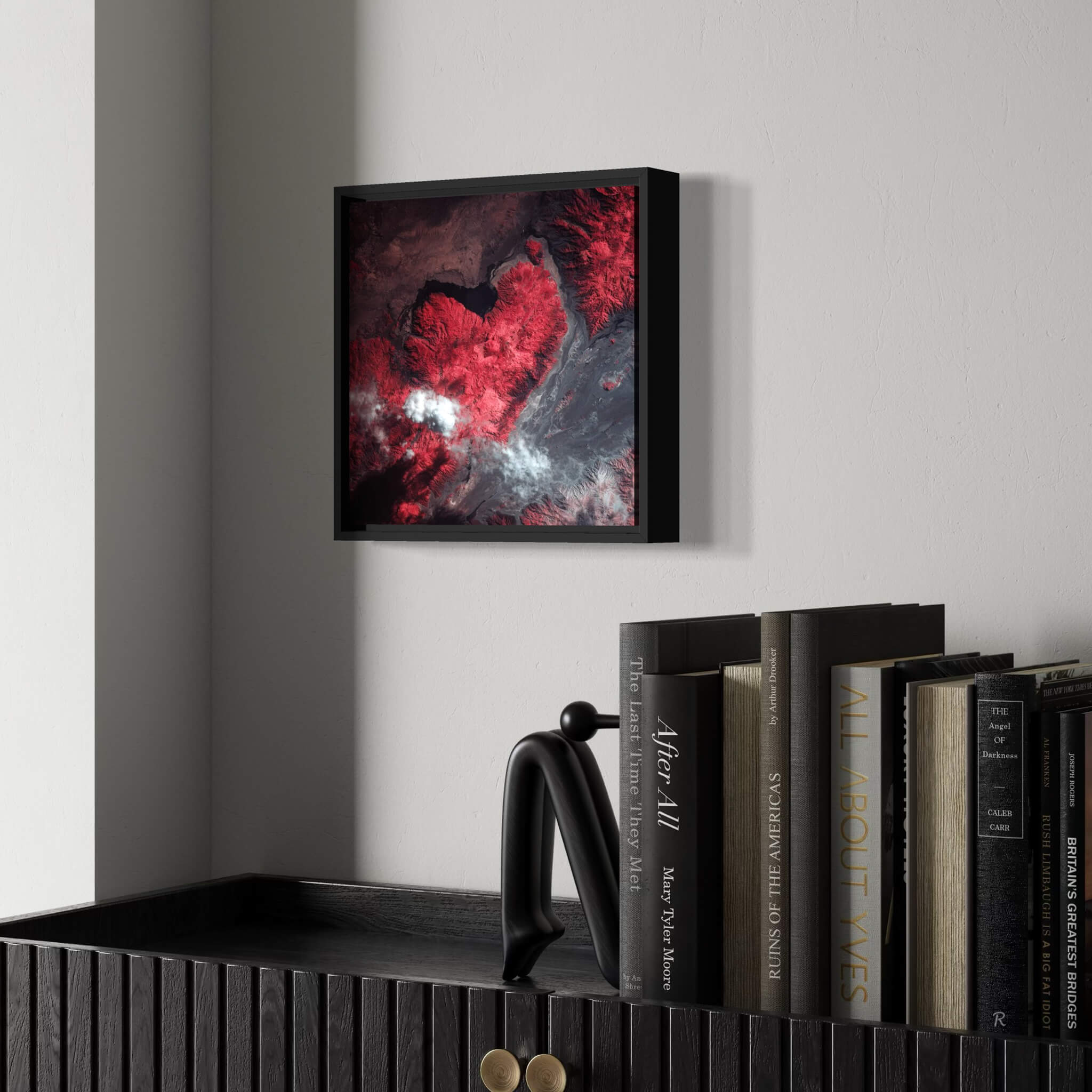 Framed artwork of an infrared satellite photography of a mountain shaped like a heart  on a wall above a bookshelf with books.