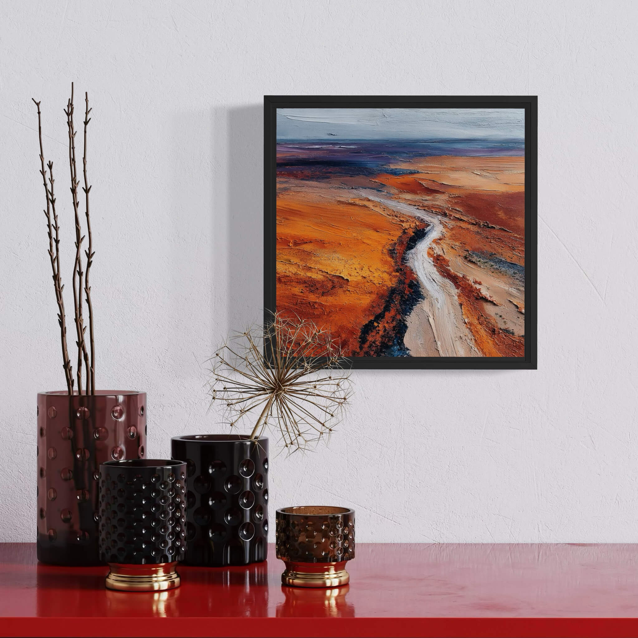 Framed landscape painting on a wall above decorative vases on a red surface.
