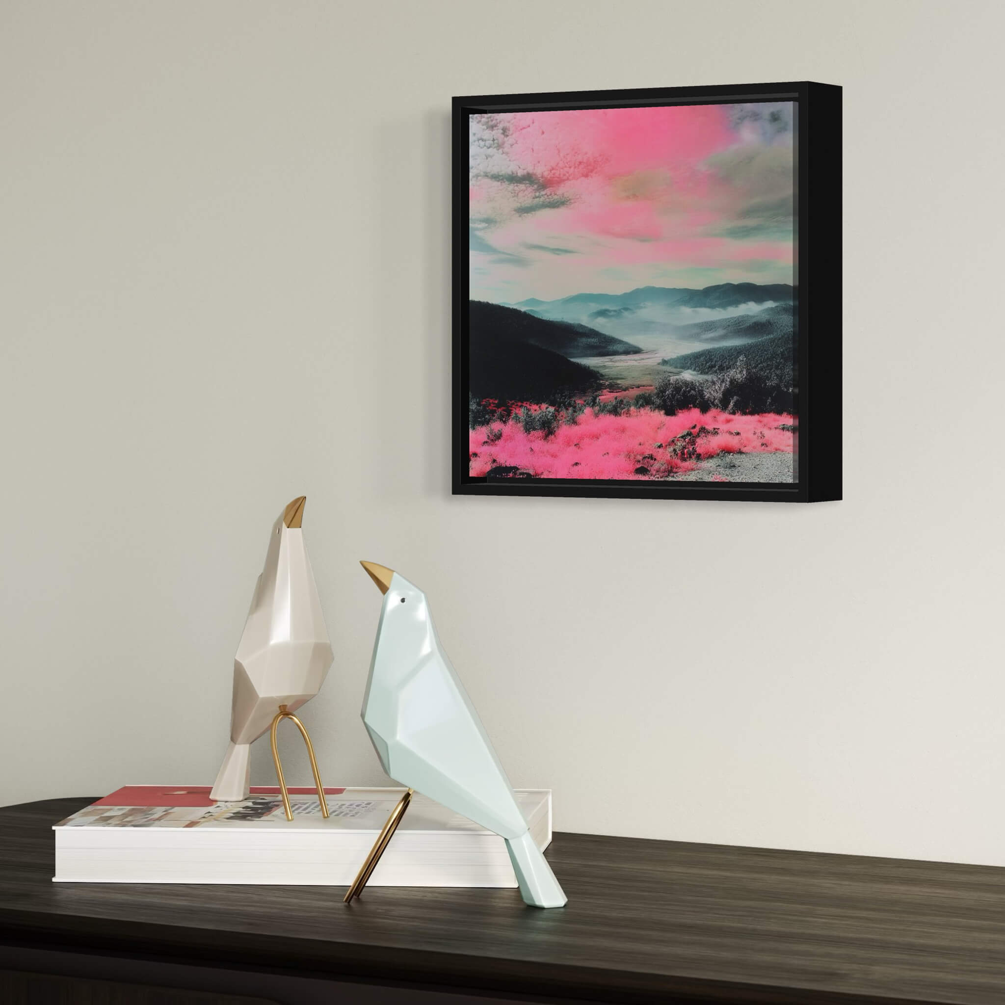 Framed abstract artwork with pink and blue hues landscape on a wall above decorative birds and books.