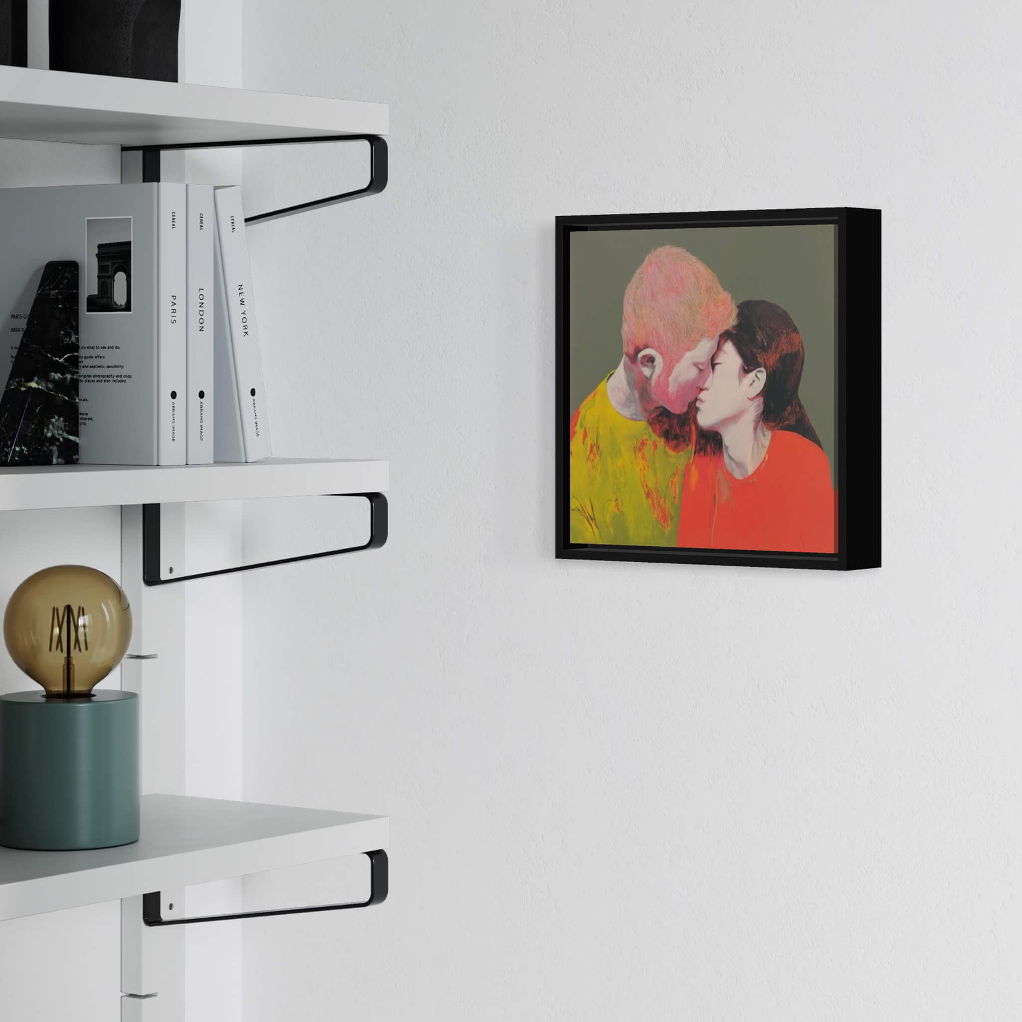 Framed artwork of two people kissing on a white wall above a shelf with books and a lamp.
