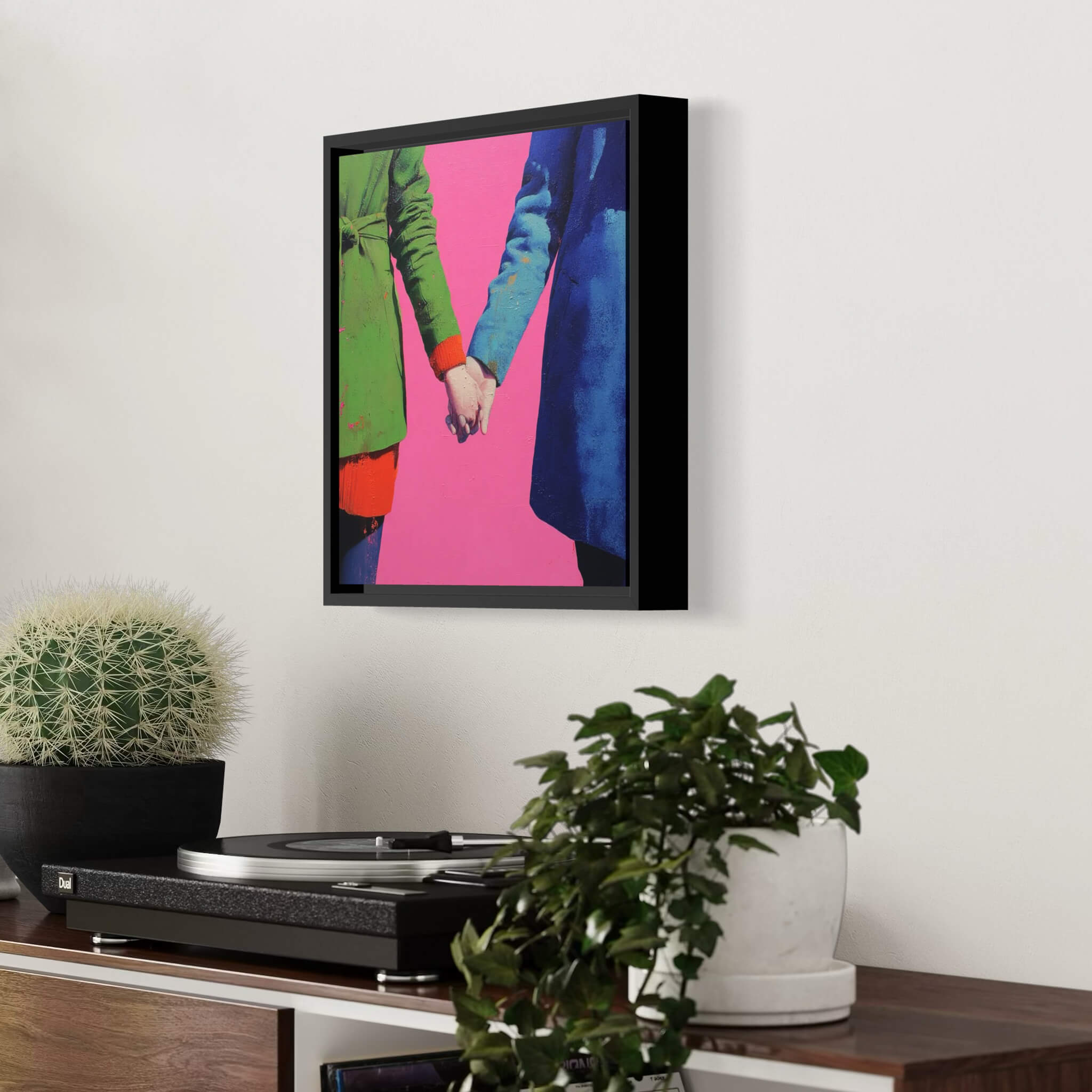 Framed artwork of two people holding hands against a pink background, displayed on a wall above a turntable.