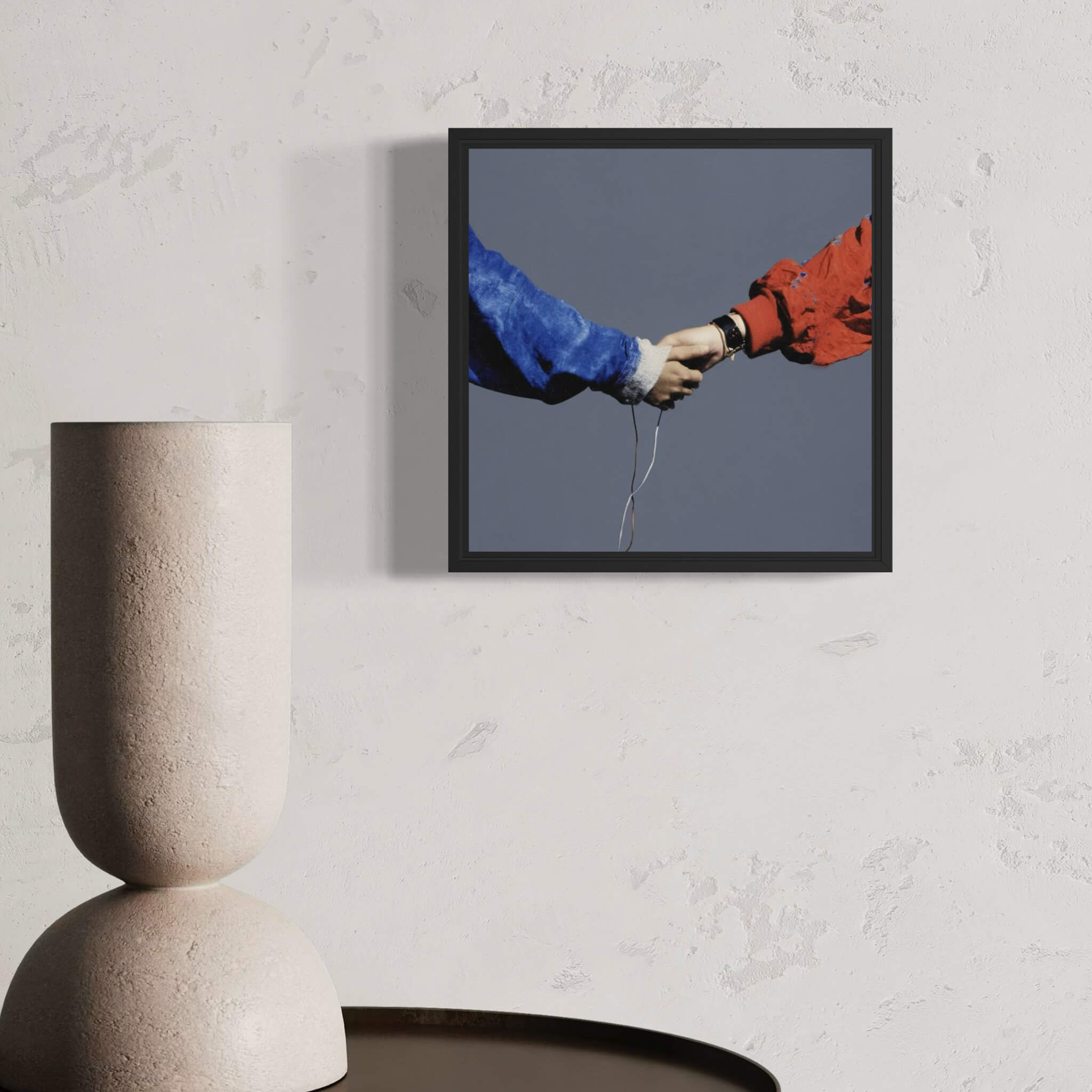 Framed artwork of two hands reaching towards each other, one in blue and one in red, on a textured wall above a modern vase.