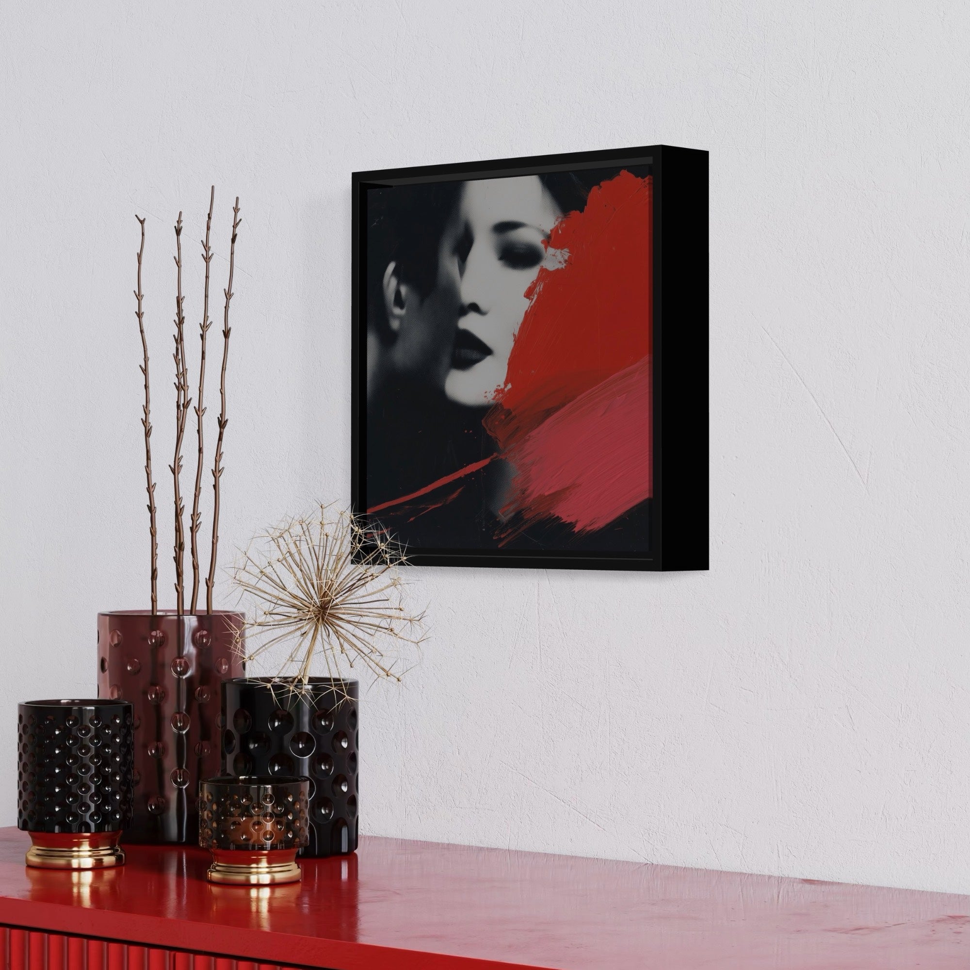 Framed artwork of a woman's face with red accent on a white wall above a red surface with decorative vases.